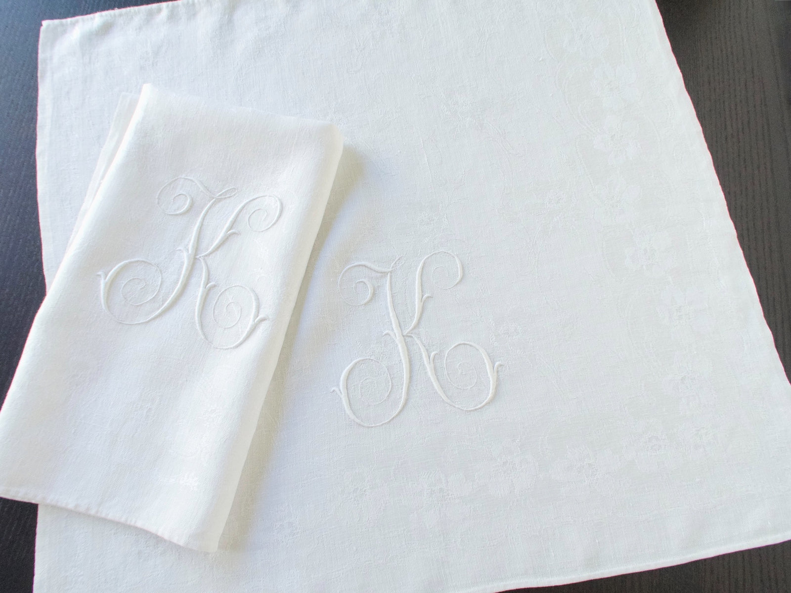 Folded and unfolded white damask linen napkins showing their large embroidered monograms.