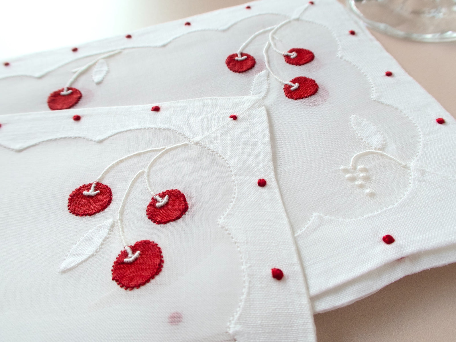Close-up detail of vintage Madeira cocktail napkin showing red cherry appliqué, white leaf embroidery, and scalloped repliqué border with red dot accents
