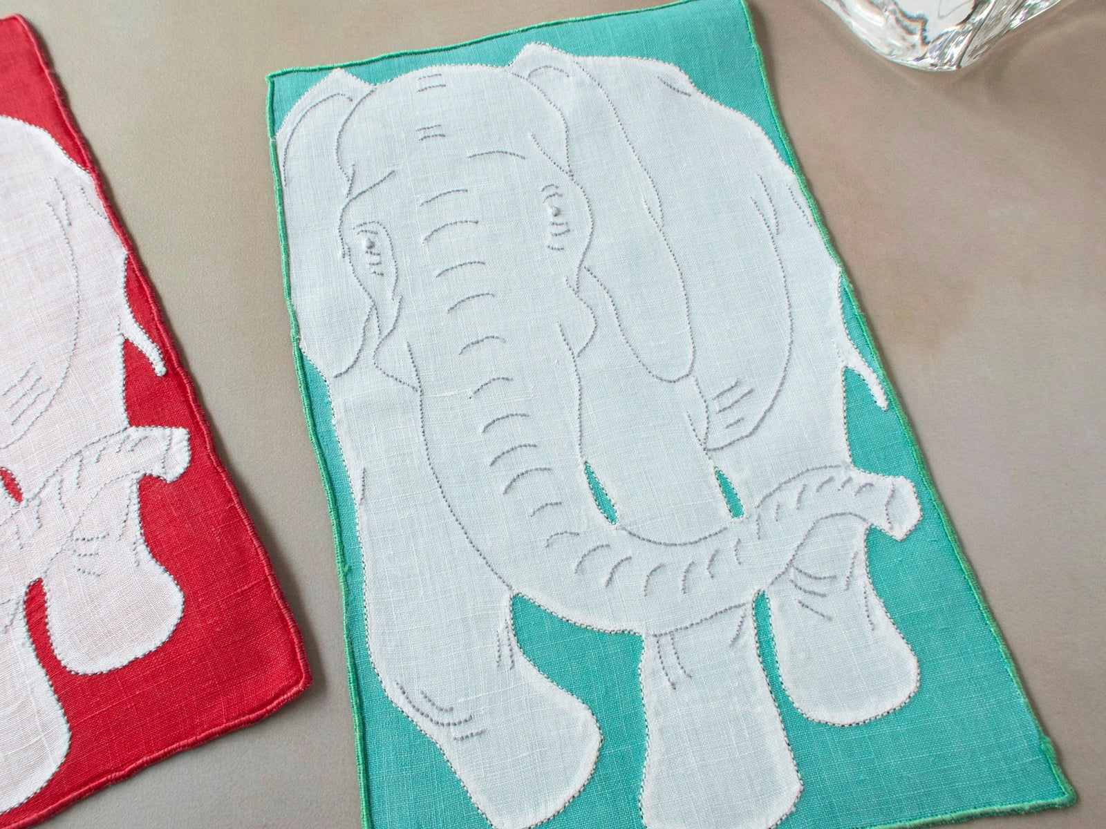 Vintage Madeira linen cocktail napkin with white elephant appliqué on teal background, with red napkin alongside on beige surface