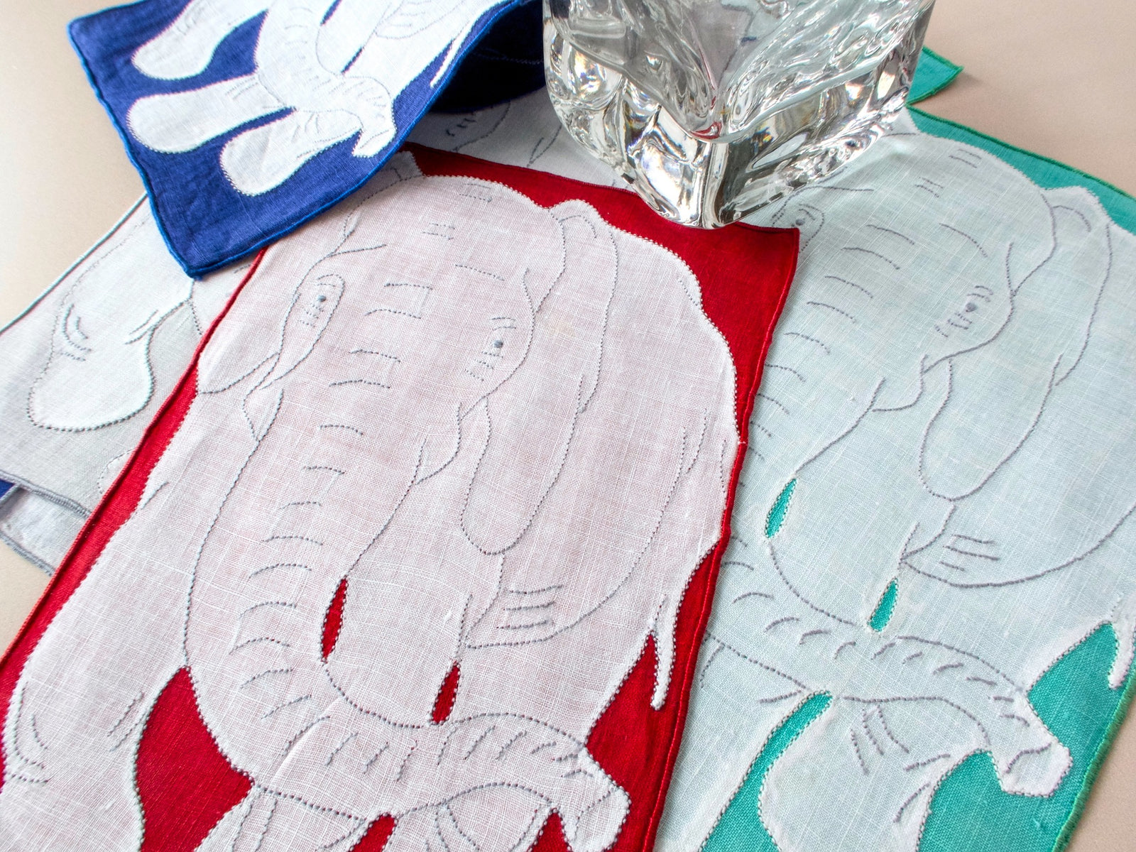 Three vintage Madeira elephant cocktail napkins in red, teal, and blue with glass for scale showing white linen appliqué detail