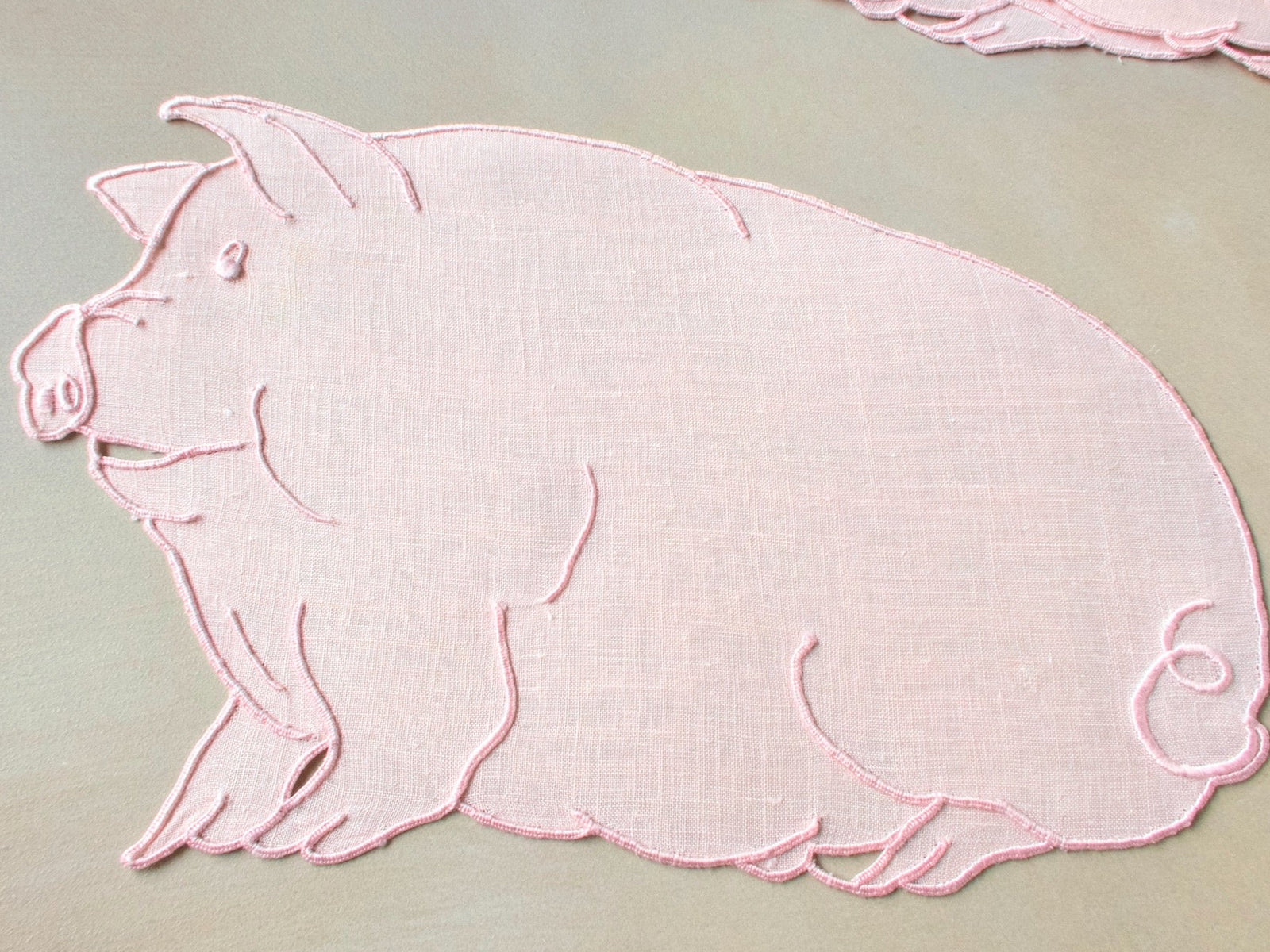 Close-up of a single pink Irish linen cocktail napkin cut and embroidered in the shape of a plump pig with curly tail, Madeira work.
