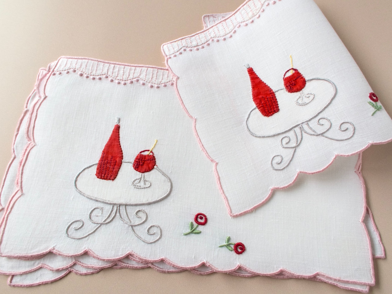 Vintage Madeira cocktail napkins fanned on beige background, red wine bottle and glass embroidered on scrollwork café table with roses, pink scalloped border.