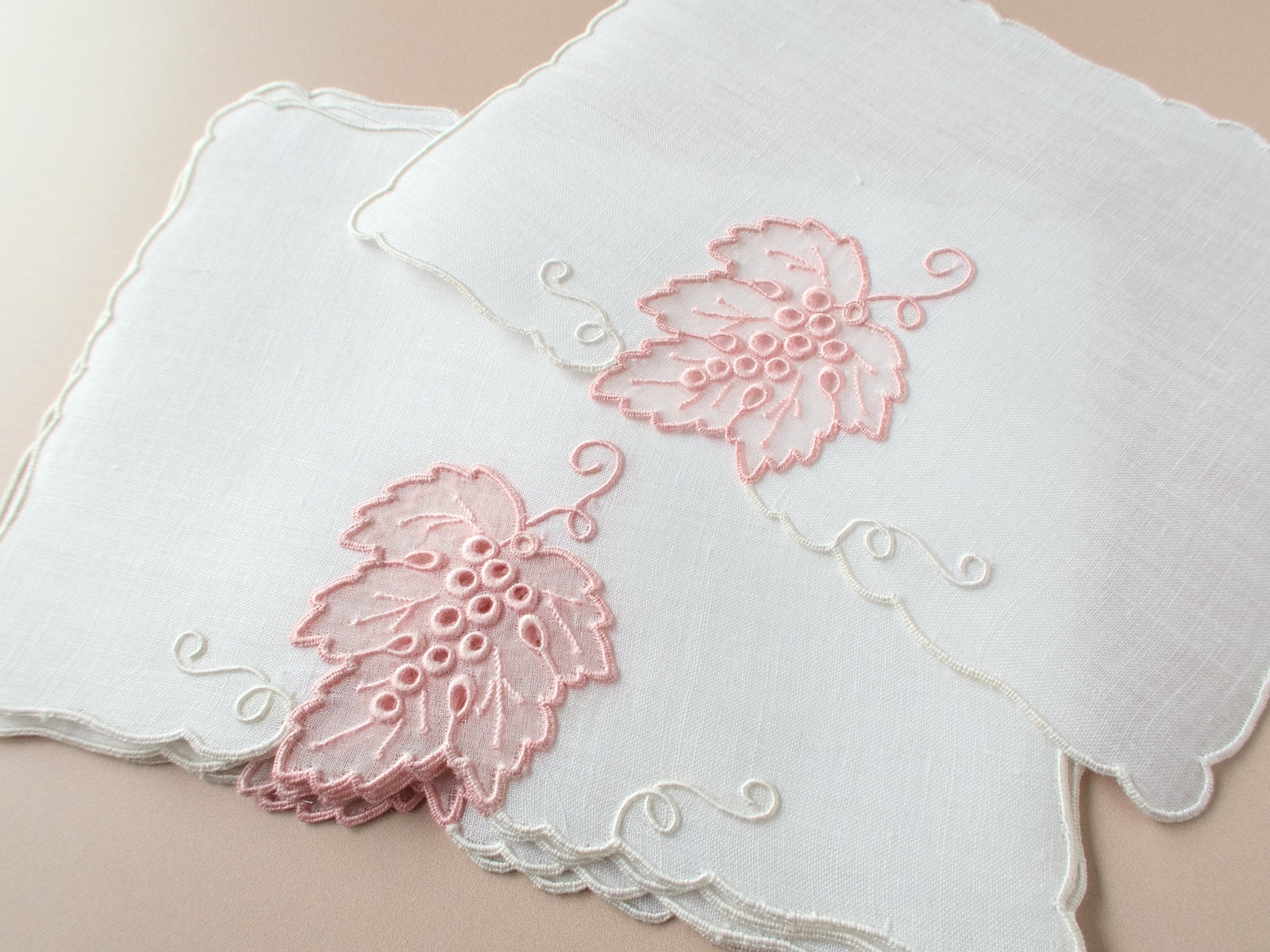 Vintage Madeira cocktail napkins on beige background, two napkins showing blush pink organdy grape leaf with embroidered veins and raised grape clusters with scrolling white vines.