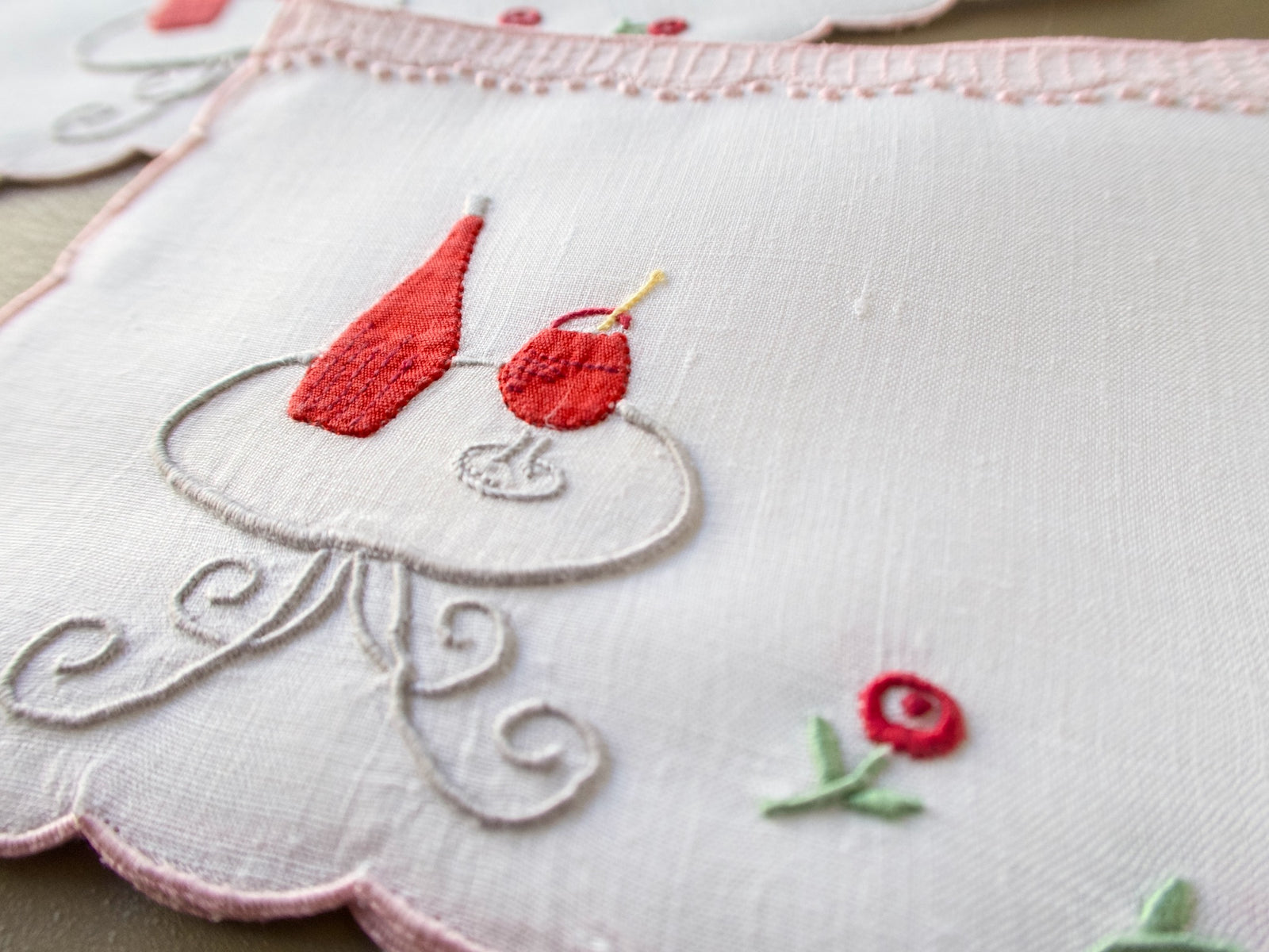 Close view of a hand embroidered bottle, glass and rose on a scrollwork café table on a vintage Madeira cocktail napkin