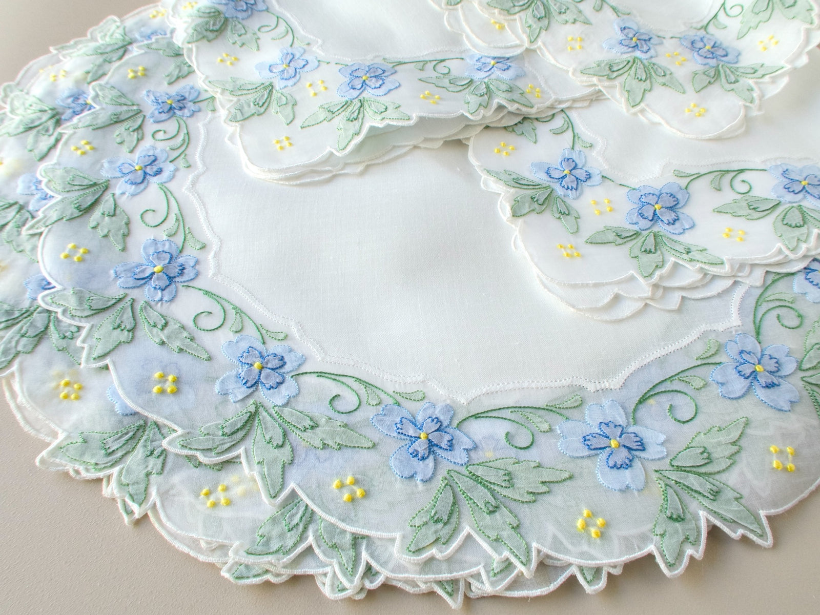 Detail of stacked vintage Madeira placemats and napkins showing hand embroidered blue flowers, green leaves, and yellow granitos on organdy