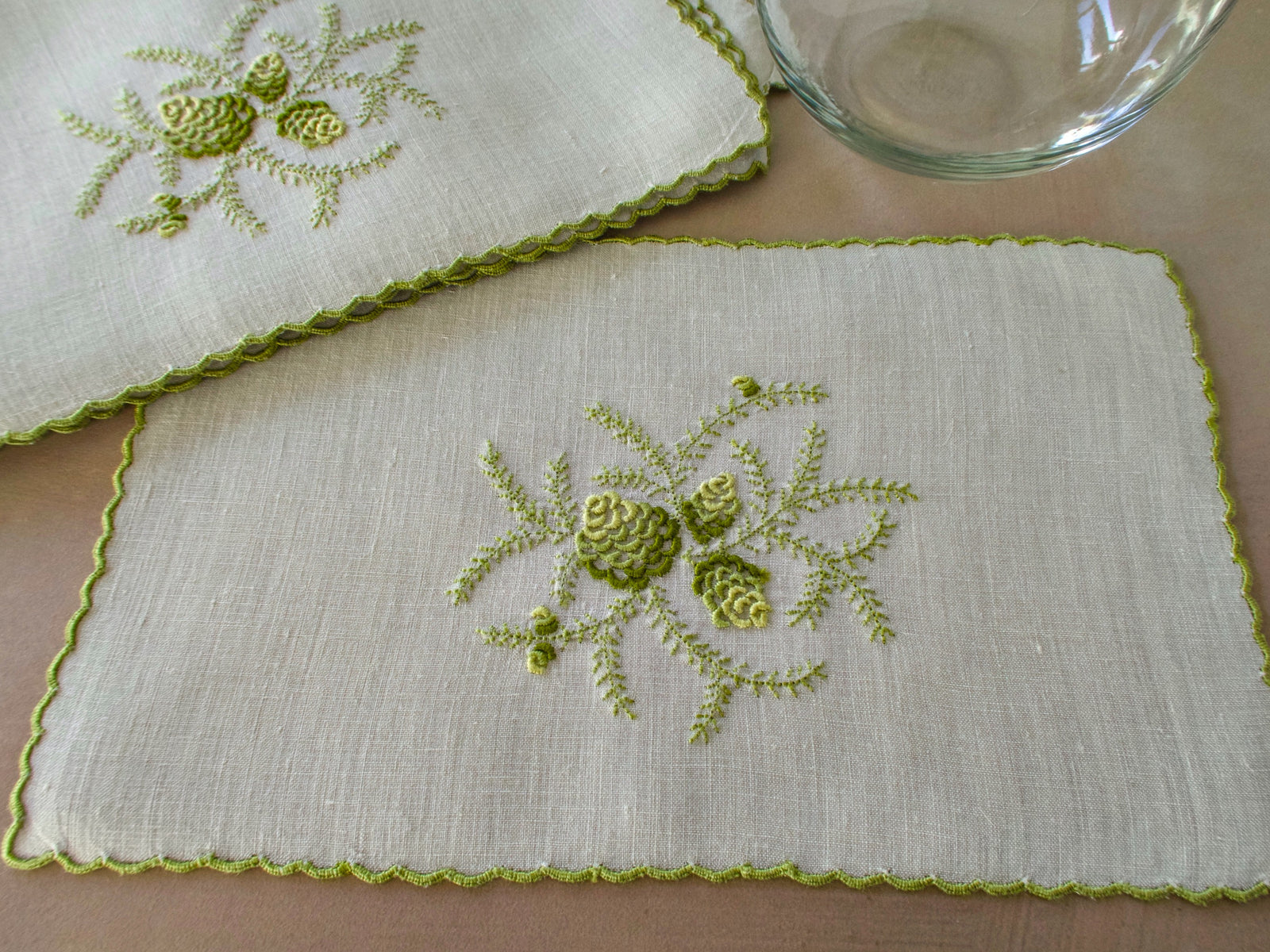 Green embroidered pinecone motif on linen cocktail napkin, scalloped edge.