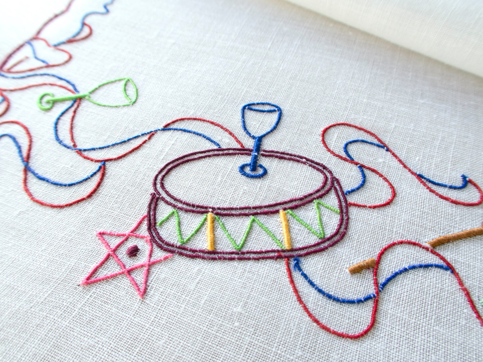 Close-up of embroidered drum & ribbon motif on white linen napkin, hand embroidered in Madeira
