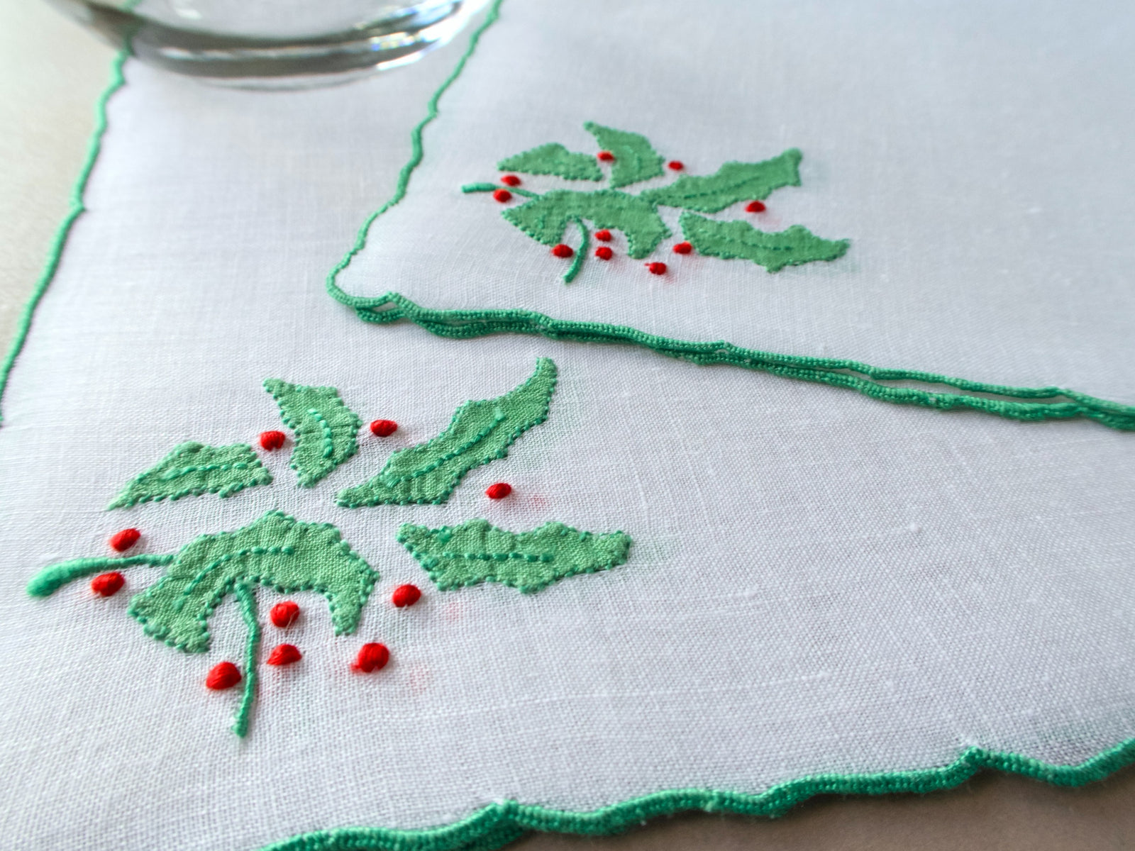 Close-up of white square linen napkin with scalloped green edge and embroidered holly leaves and red berries.