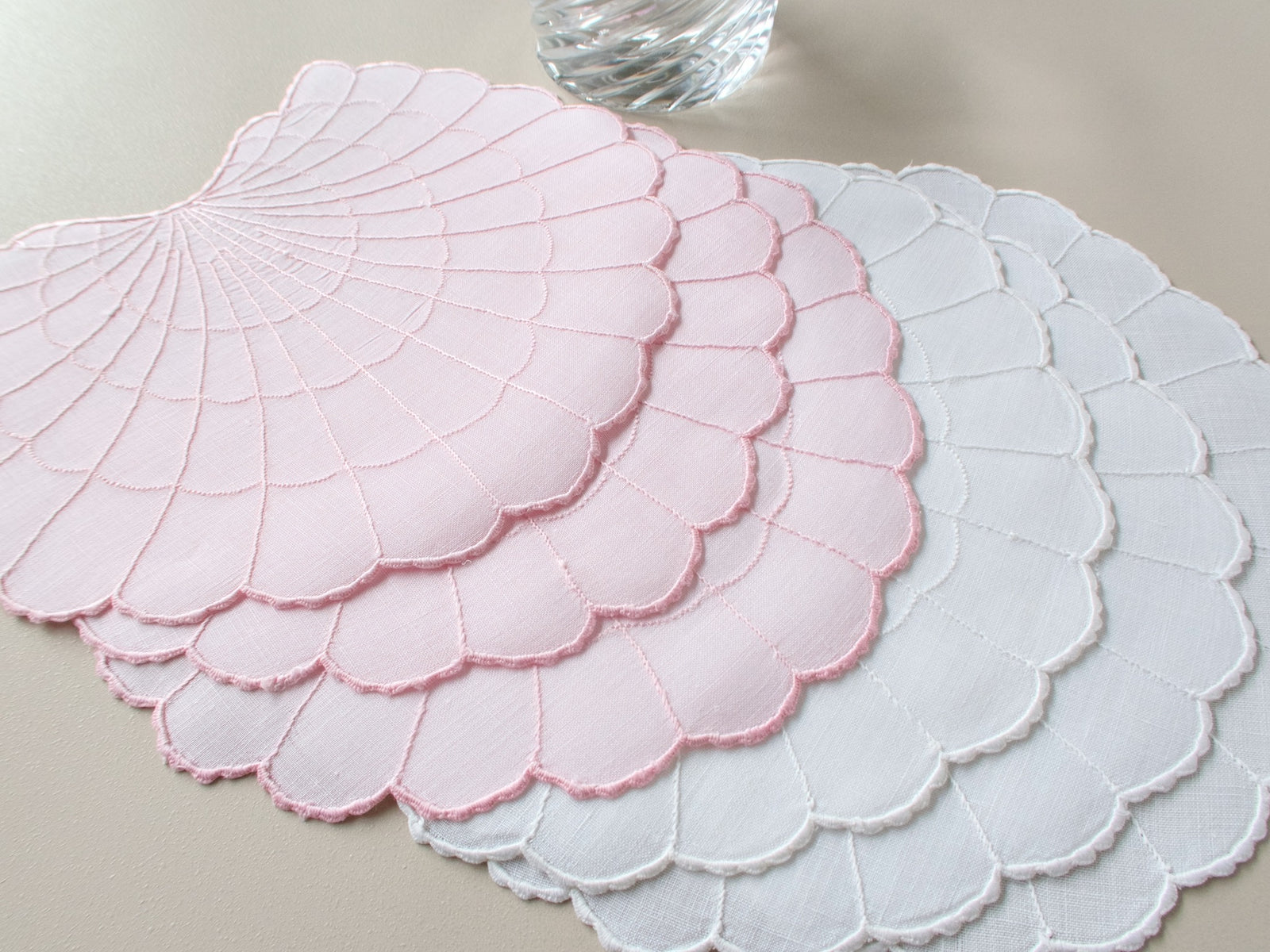 Pink and white scallop shell-shaped Madeira cocktail napkins, with radiating embroidered lines and scalloped edges, layered on a warm taupe background.