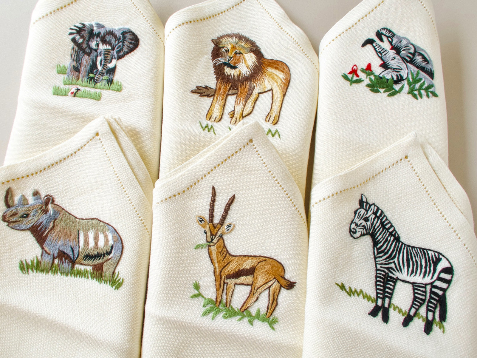 Six vintage hand embroidered safari napkins folded showing full set including zebra, lion, gazelle and elephant designs
