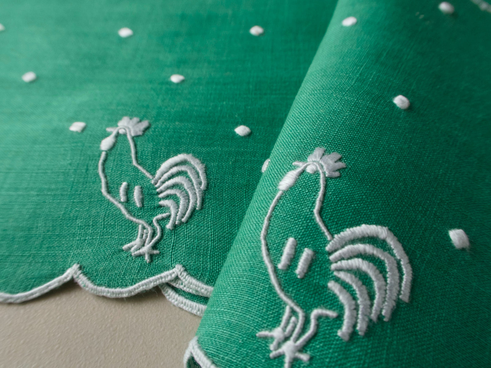 Green linen cocktail napkins with white embroidered rooster and snowy dot motif, folded to show scalloped edge.