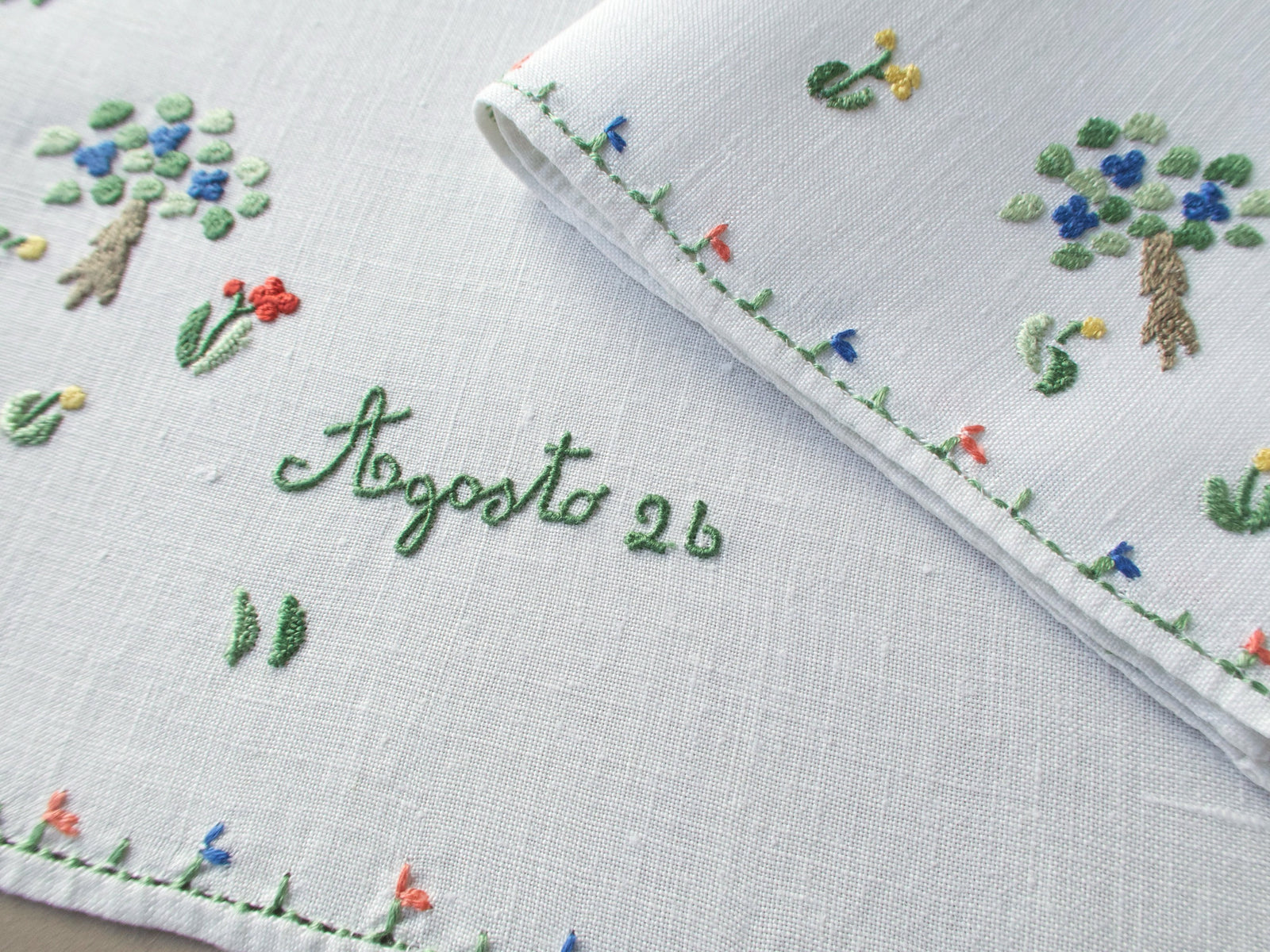 Close-up of the embroidered inscription “Agosto 26” framed by tiny flowers and foliage on white linen.