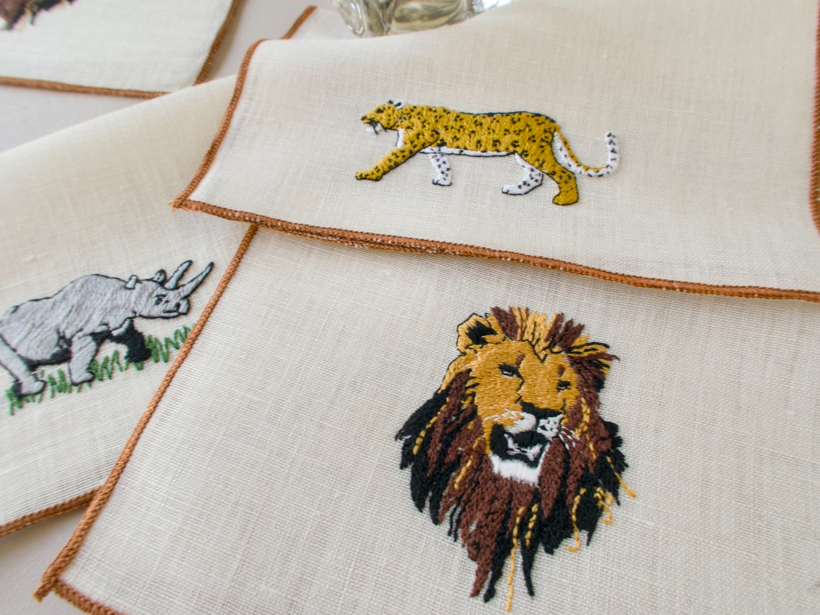 Three vintage cocktail napkins fanned out on ecru linen with brown border, showing cheetah, rhino and lion embroidery