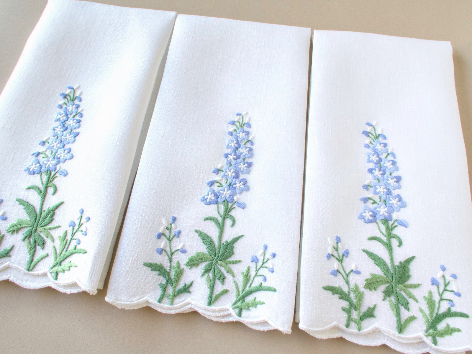 Three white Madeira linen guest towels displayed side by side, each hand embroidered with a tall delphinium spike in periwinkle blue and white above spreading green foliage, scalloped lower hem