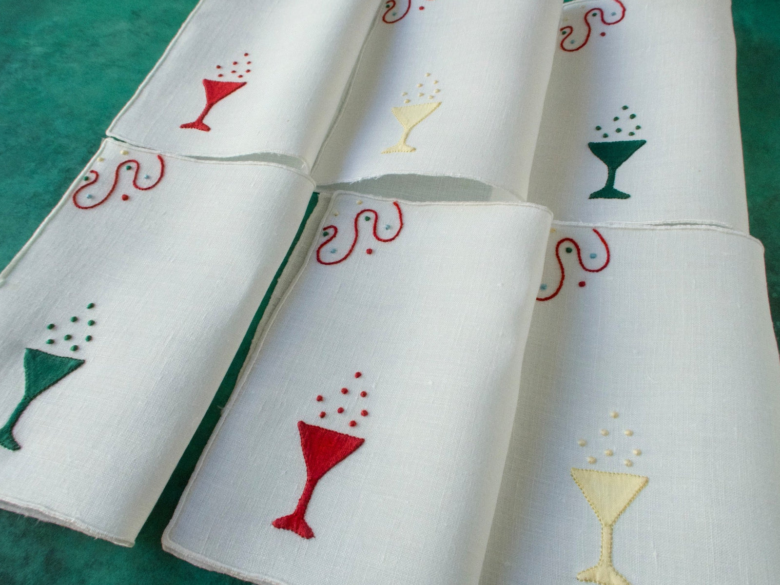 White linen cocktail napkins embroidered with champagne glasses in red, green and yellow, arranged in a group.