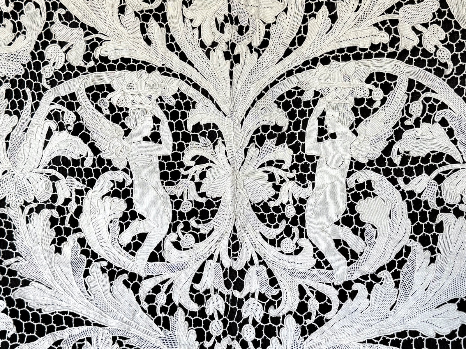 Two winged figures bearing baskets amid flowers and scrolling leaves in the lace.
