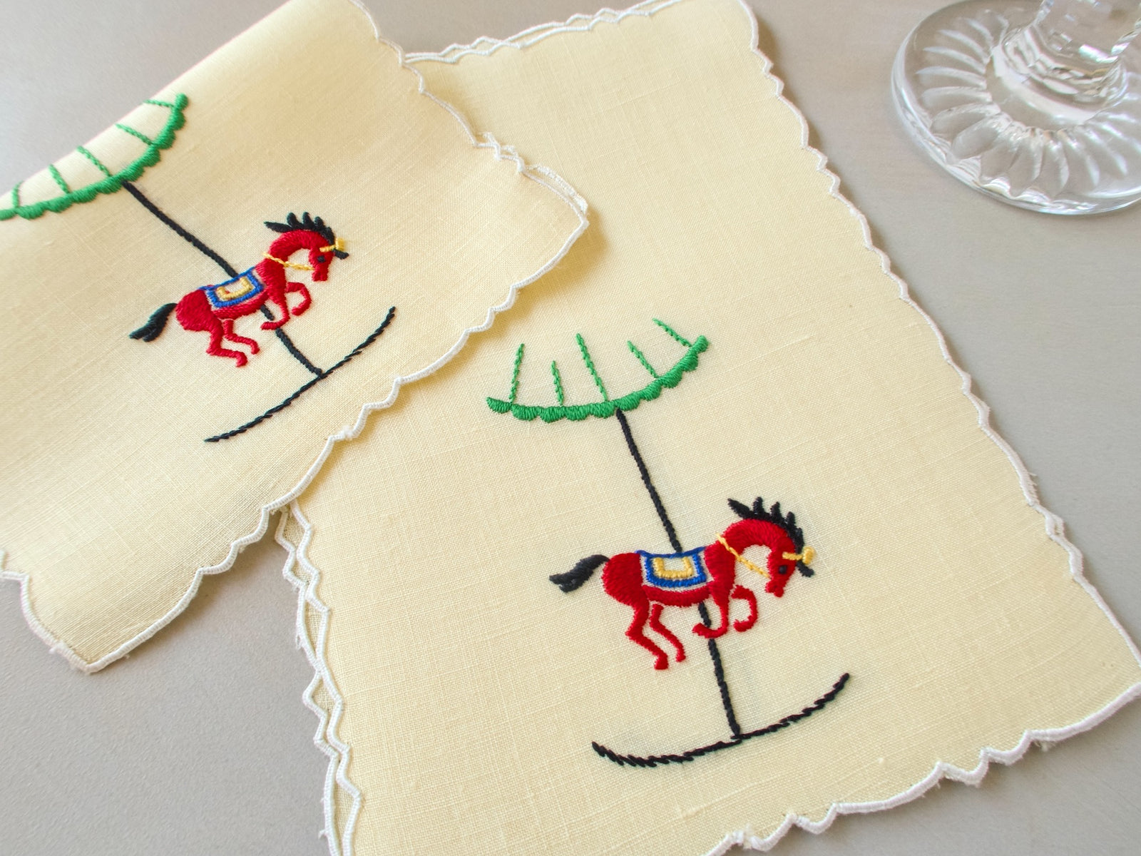 Stacked vintage Madeira cocktail napkins, yellow linen with scalloped edges, embroidered red carousel horse with blue and gold saddle on a black pole
