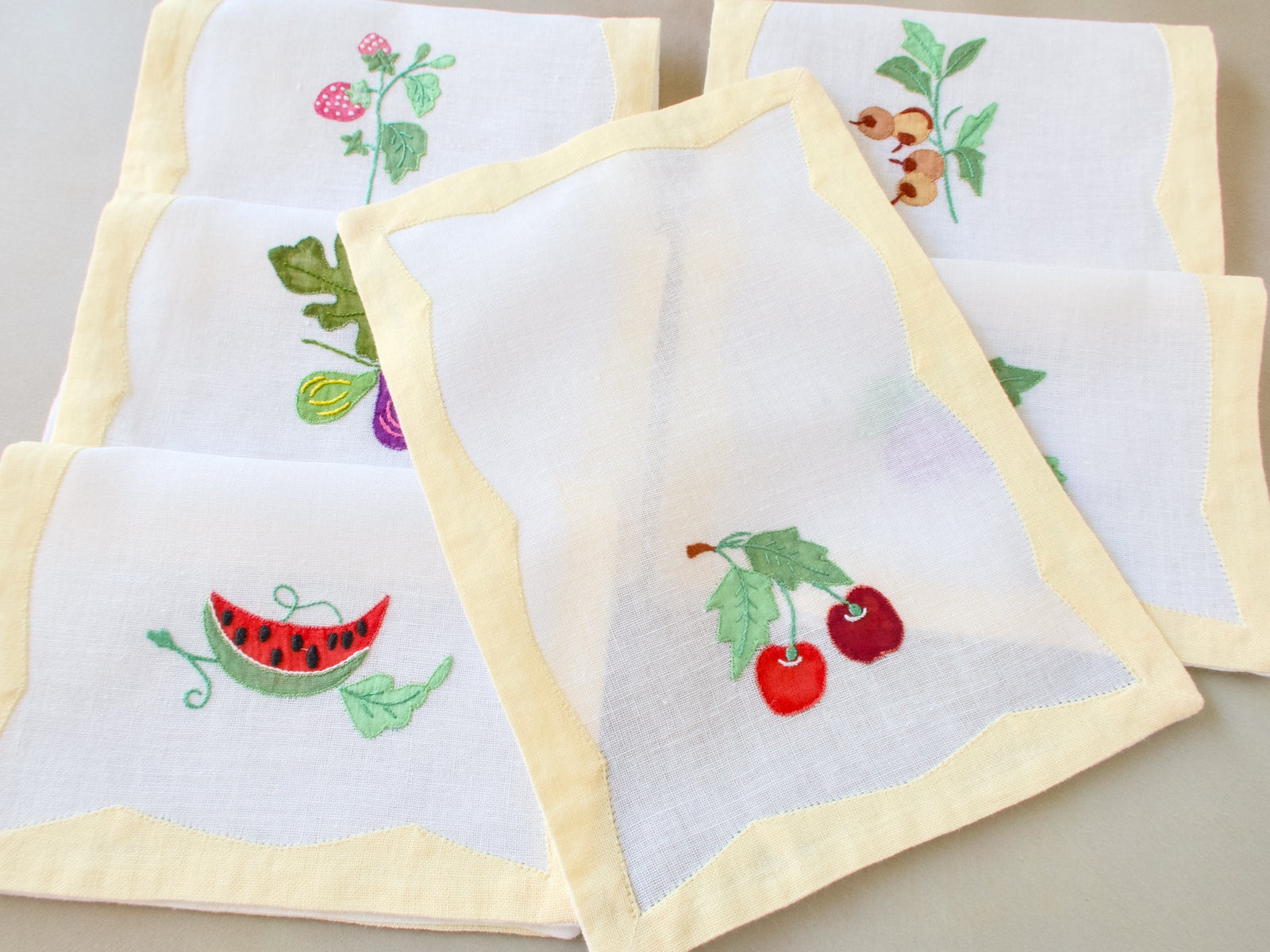 Single vintage Italian cocktail napkin unfolded over the set, showing semi-sheer white linen quality with yellow border and cherry embroidery, five napkins visible beneath