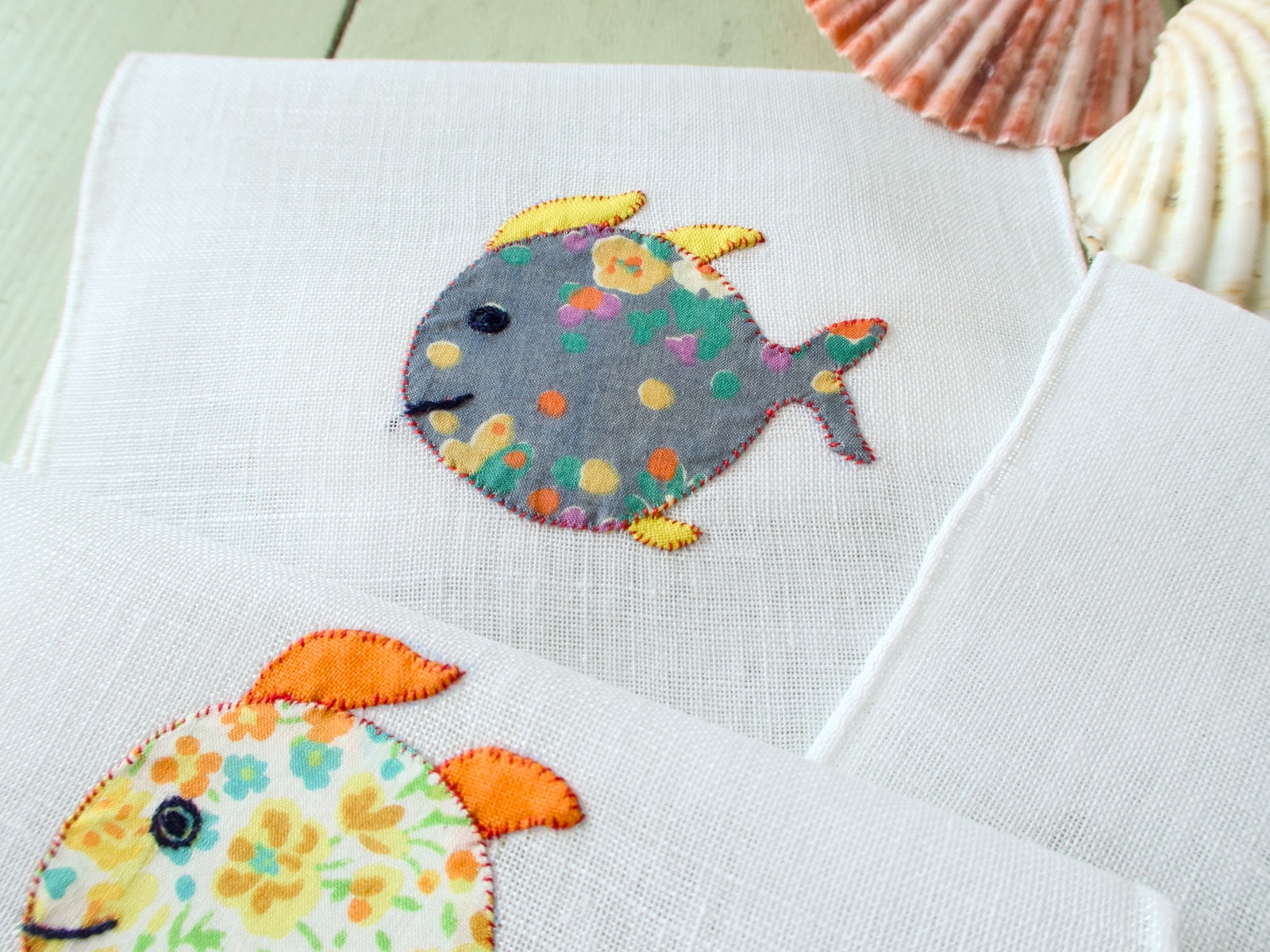 Close-up of two vintage Italian cocktail napkins, grey floral and yellow floral appliqué fish detail on white linen