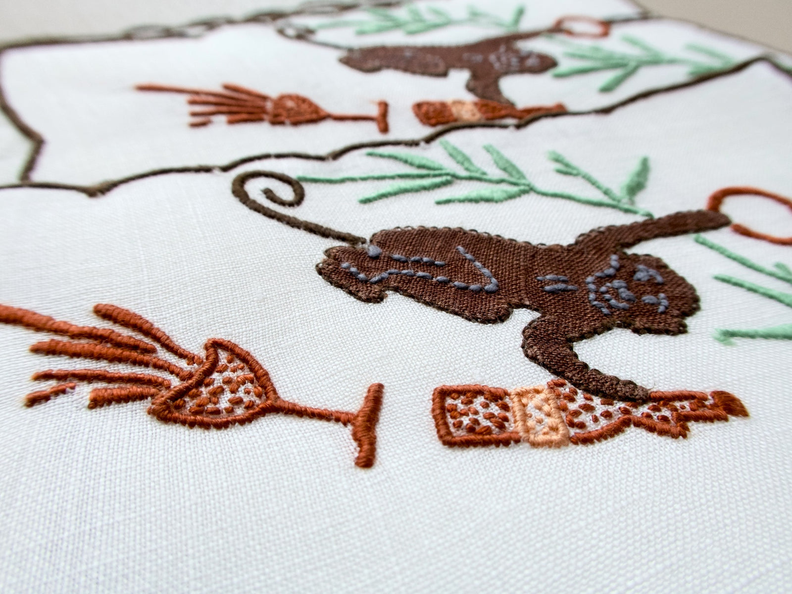 Close-up of vintage Madeira cocktail napkin showing brown appliqué monkey swinging sideways, with spilled champagne glass