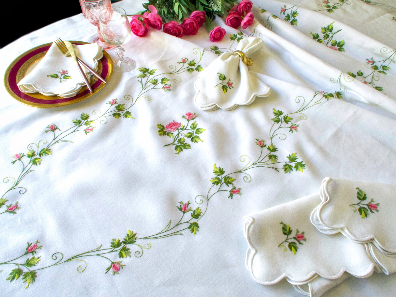 D. Porthault oval tablecloth on round table set with gold-rimmed burgundy charger, pink crystal stemware and scalloped napkins with rosebud embroidery
