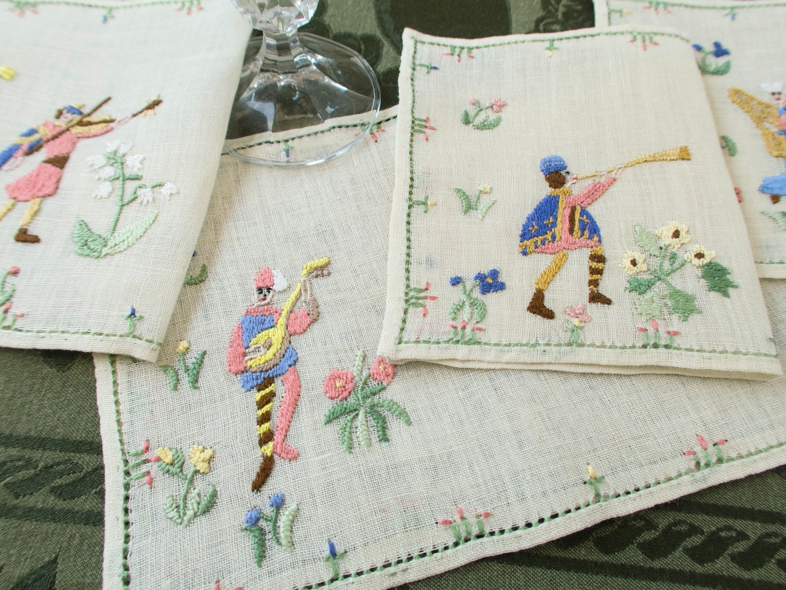 Close-up of Rapisardi medieval musician cocktail napkins with two figures and floral embroidery under a glass stem.