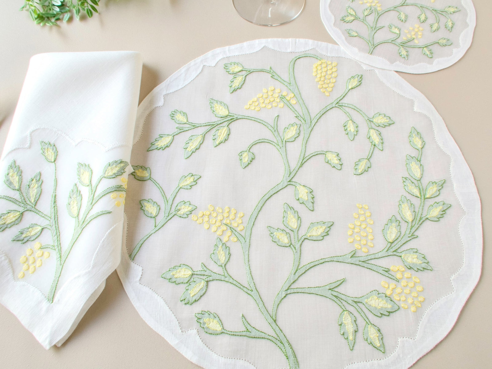 Overhead view of vintage Madeira organdy placemat with matching shadow work embroidered napkin and coaster, greenery prop on beige background