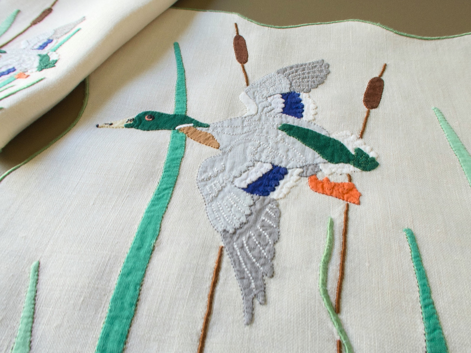 Close view of a hand embroidered mallard drake in flight among cattails on a vintage Madeira linen placemat