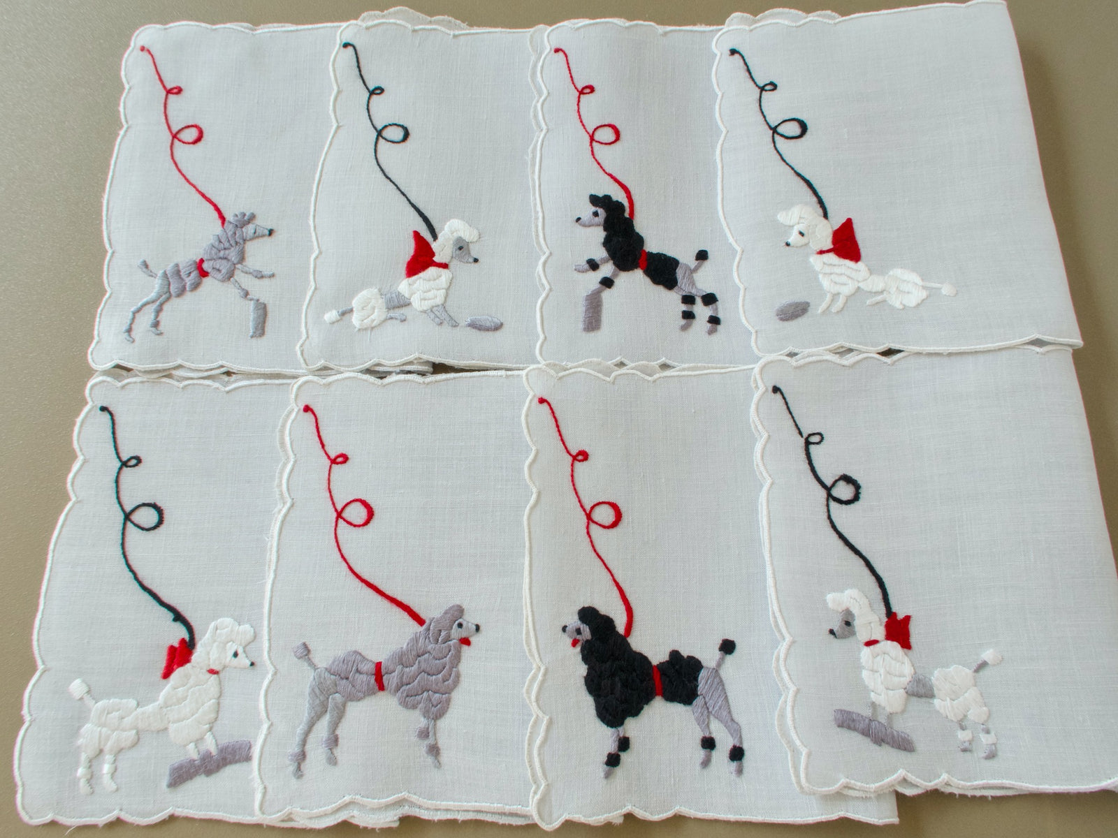 Eight vintage Madeira cocktail napkins laid out in two rows, each with a hand embroidered poodle on a looping leash