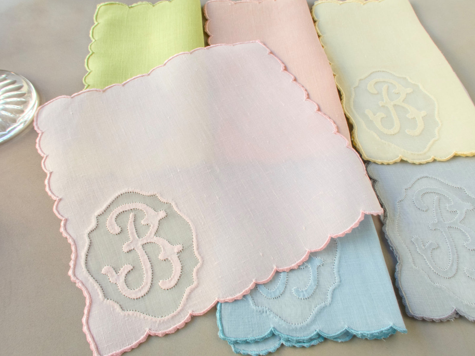 Single vintage Madeira Irish linen cocktail napkin hand embroidered with cursive “B” monogram in organdy window and scalloped edging, in pale pastel linen