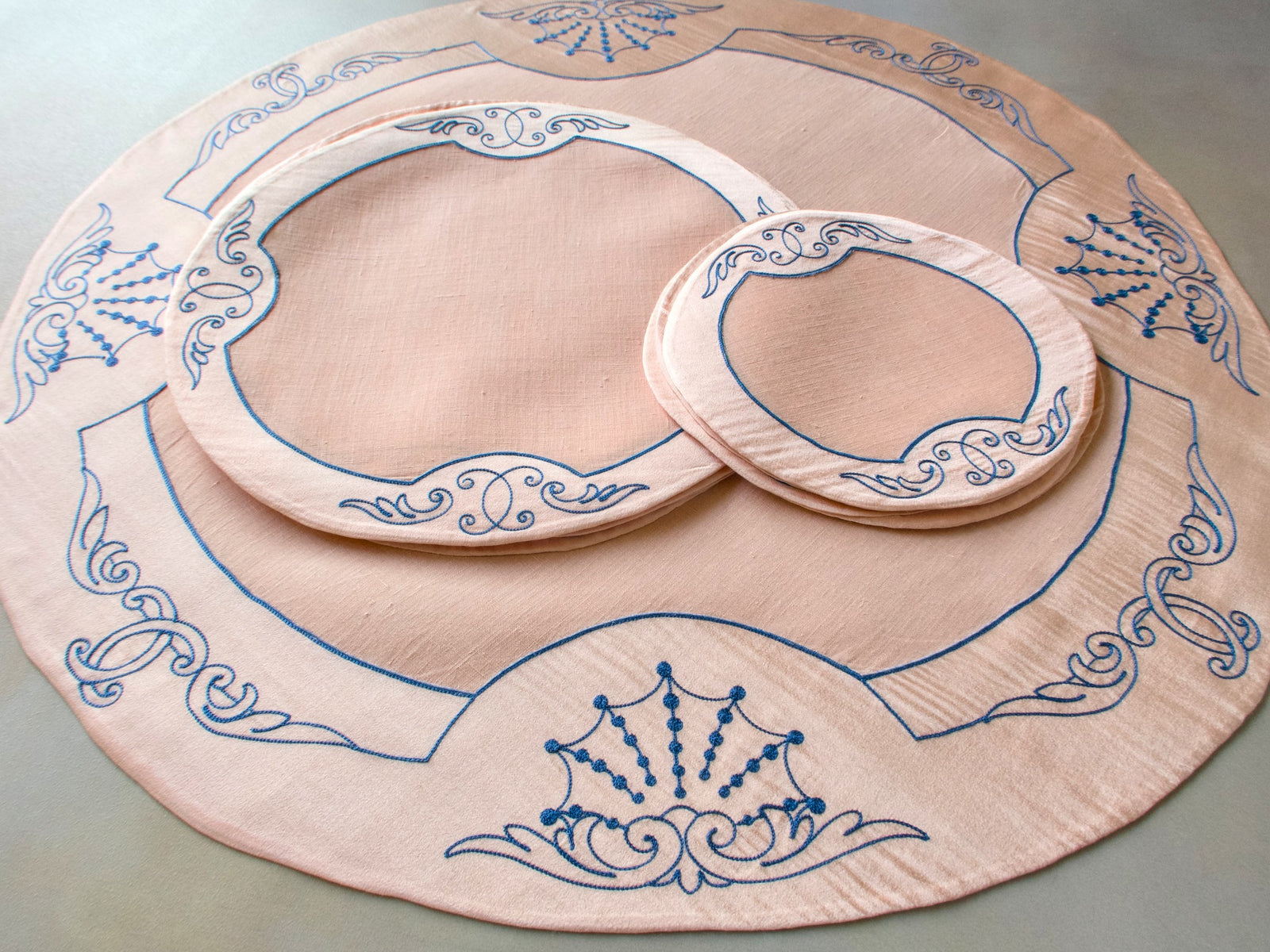 Full vintage French Cornely tea dessert set for 6 laid out, centerpiece with round placemat and coasters showing blue scrollwork embroidery on salmon linen