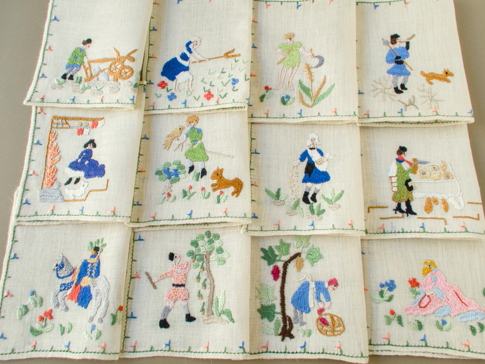 Set of 12 folded vintage Baroness Rapisardi Months of the Year cocktail napkins in three rows, each hand embroidered with a different seasonal scene from plowing and haying to the grape harvest and winter hunt, using the punto stuoia technique