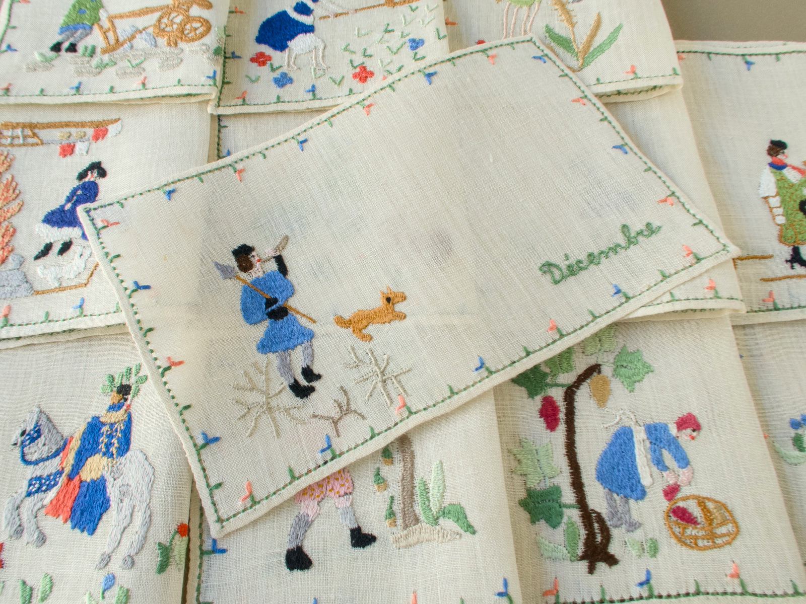 Baroness Rapisardi Décembre cocktail napkin opened flat to show a hunter in blue with a spear and horn, a small tan dog and frosted branches, with the month name in green script at right