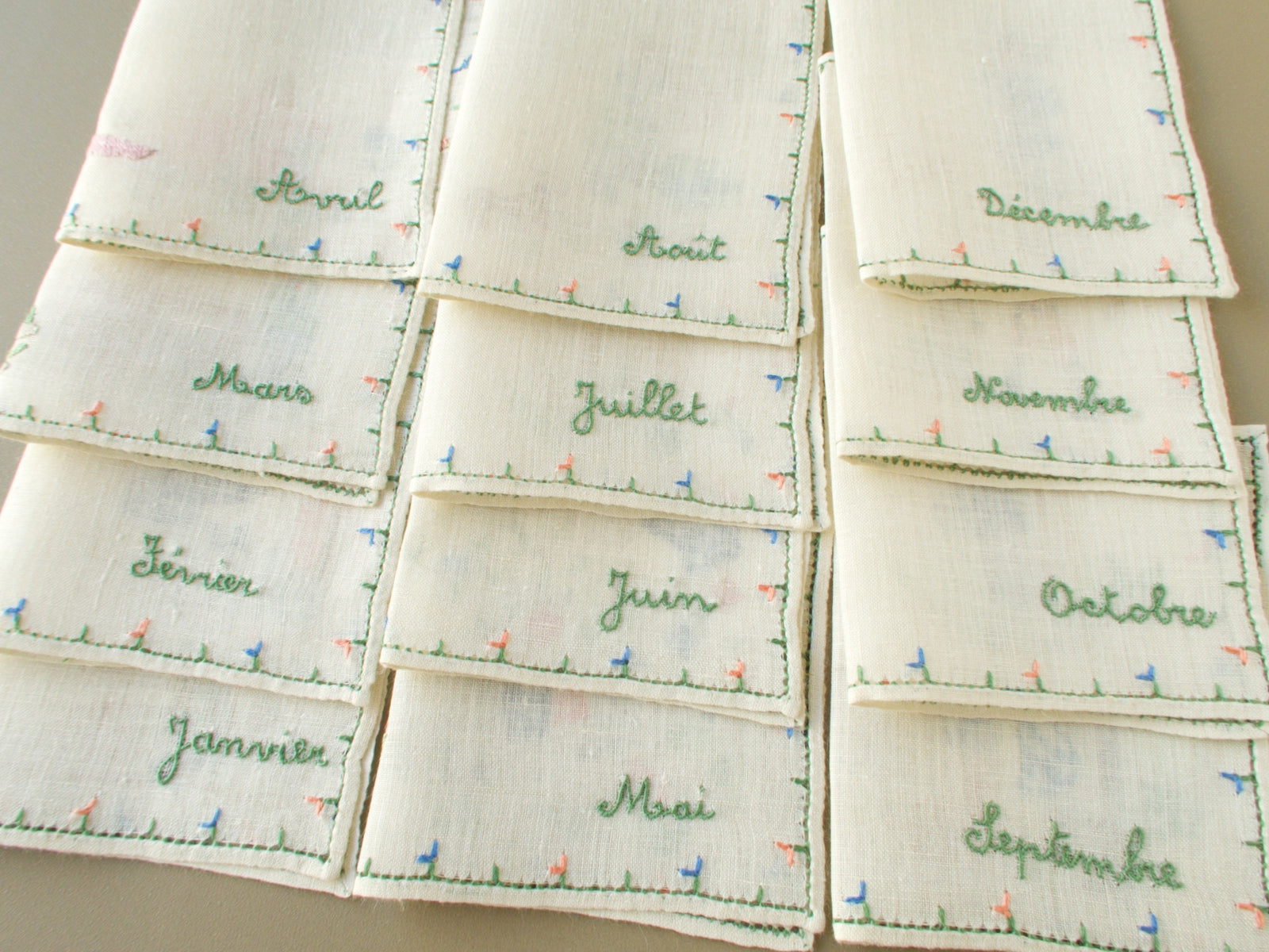 All 12 Baroness Rapisardi cocktail napkins folded to show the month names from Janvier to Décembre embroidered in green script above small pink and blue flower borders