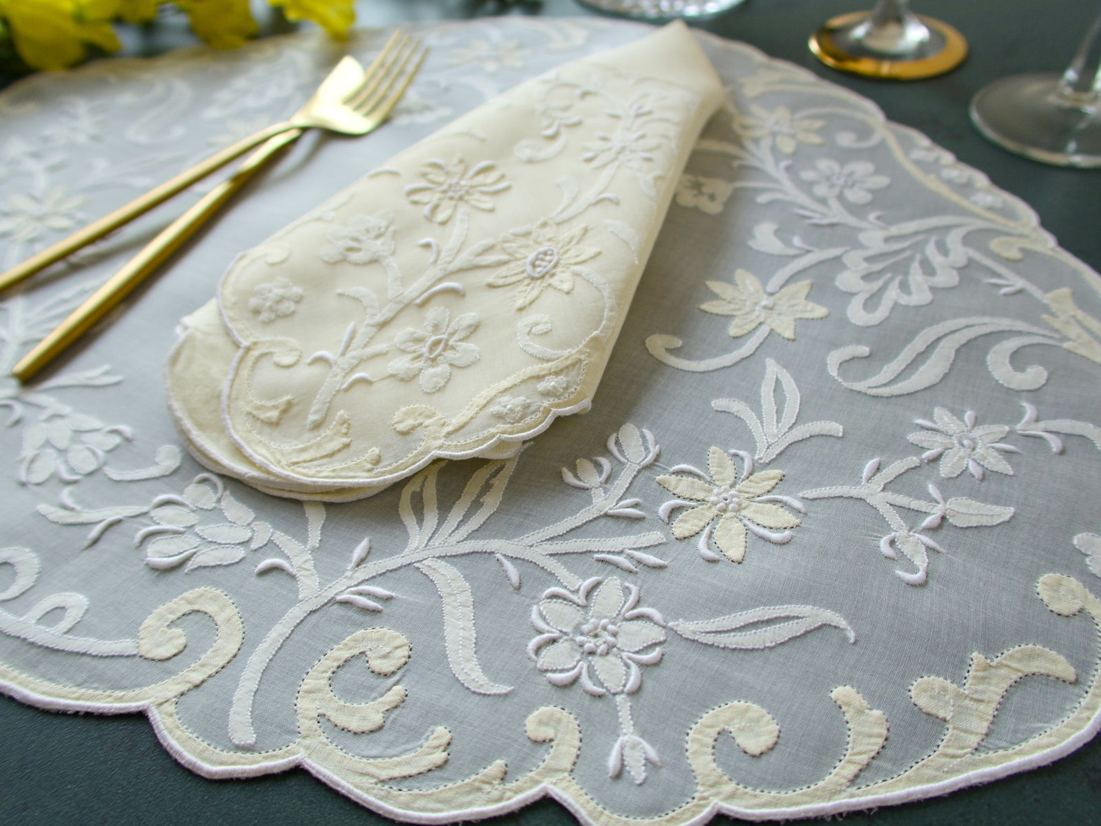 A set of vintage Madeira hand embroidered pale yellow placemats with floral embroidery, displayed on a table with cutlery and a glass.