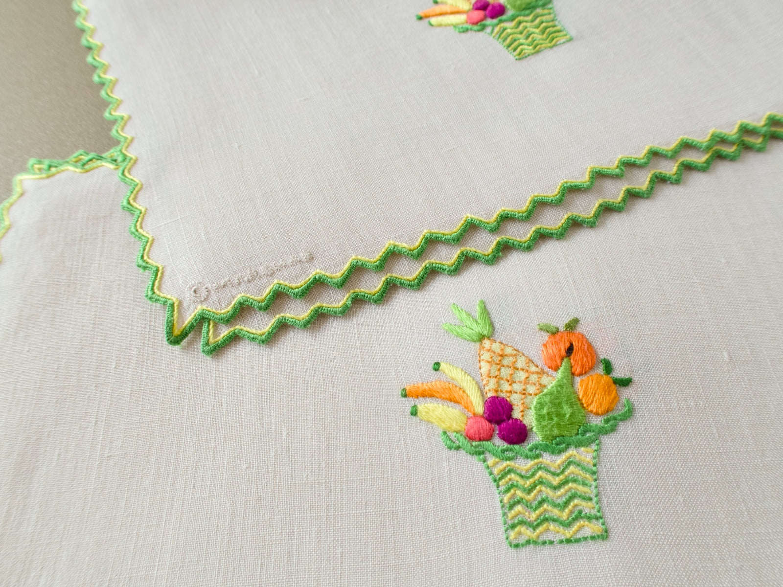 Close view of a vintage Marghab “Fruit Basket” linen cocktail napkin showing the embroidered Marghab signature, detailed fruit basket and double-stitched hem treatment.