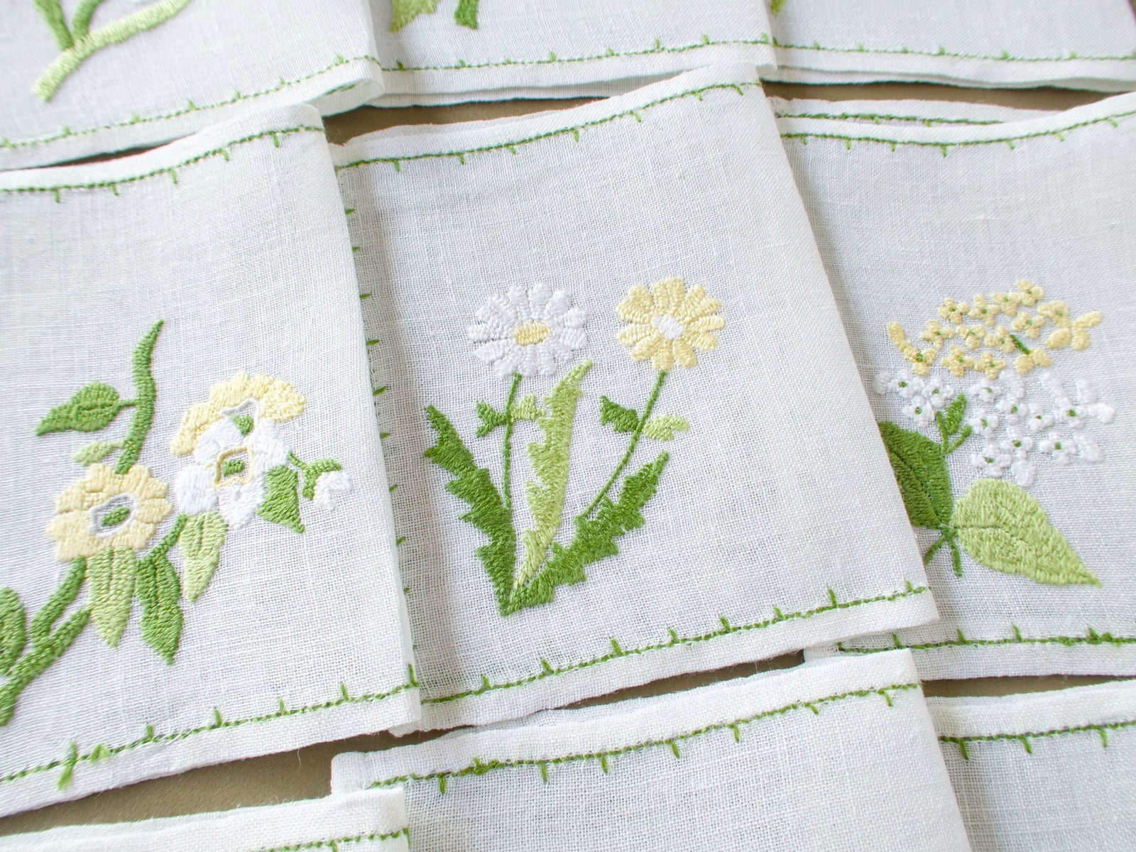 Three vintage Rapisardi cocktail napkins showing white and yellow flowers, green foliage and fine hand embroidery on semi-sheer linen.