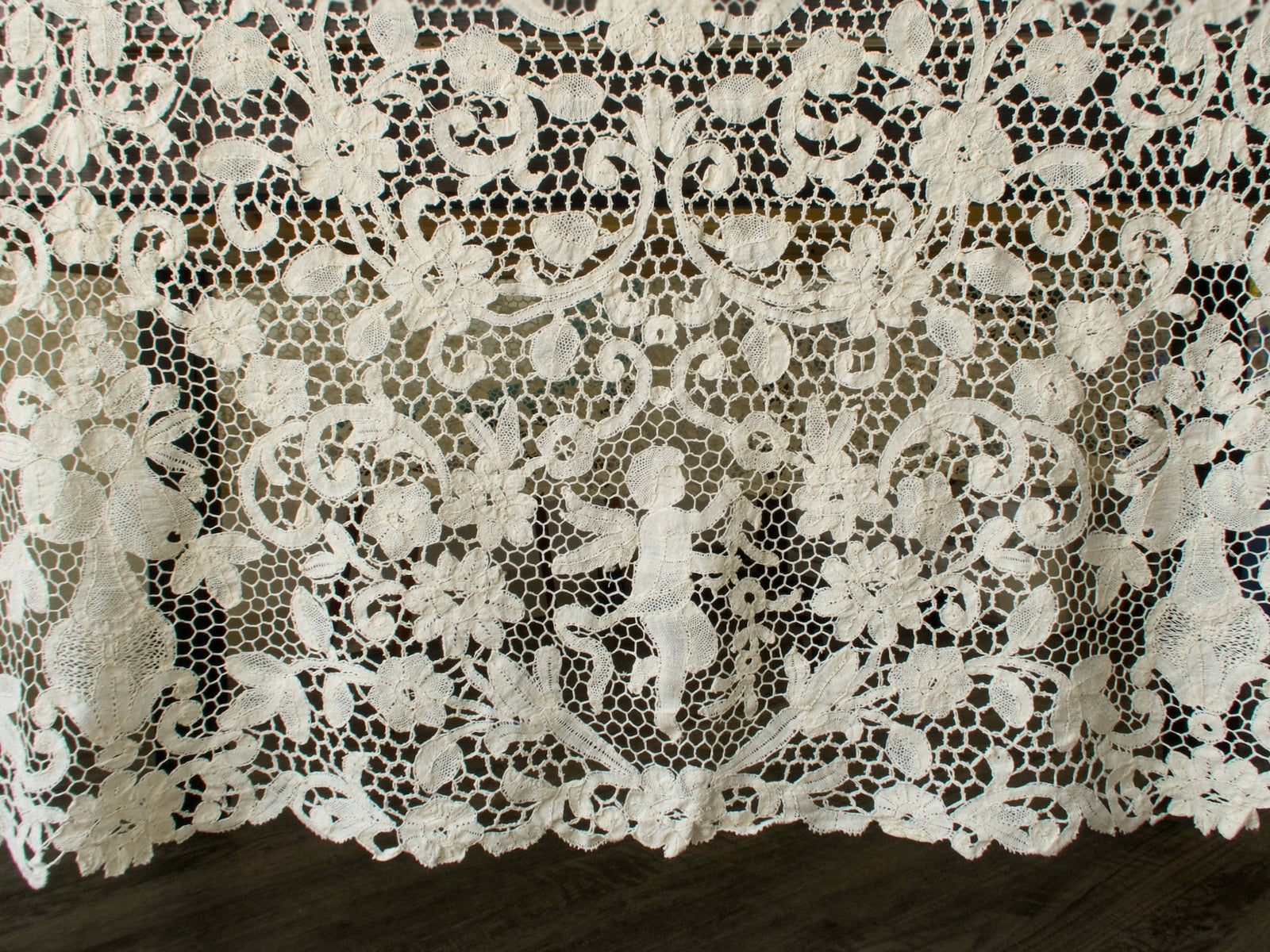 Close-up of handmade bobbin lace tablecloth showing cherubs, fruit, and floral motifs in warm ivory thread.
