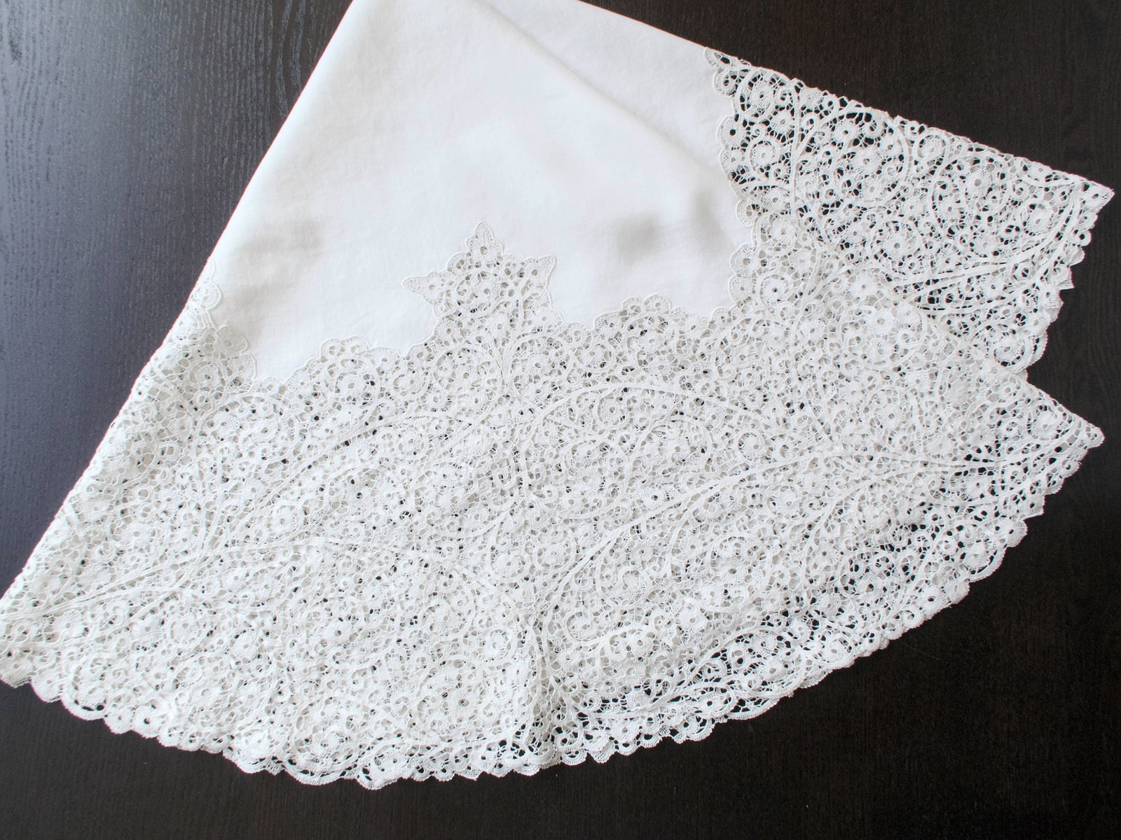 Round Italian Cantù lace tablecloth draped against a dark background, showing its linen center and wide lace border.