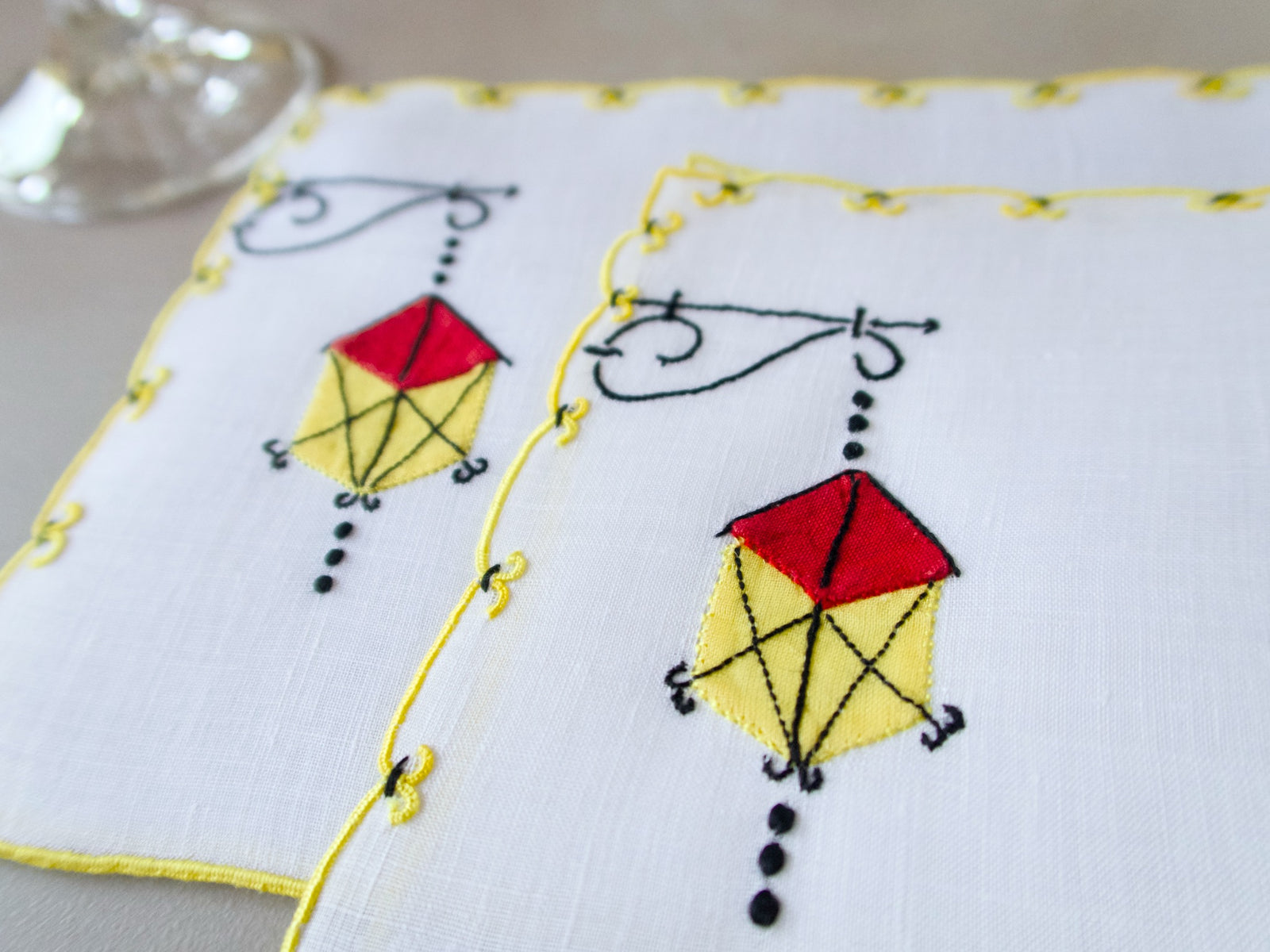 Marghab Lantern cocktail napkins, hand embroidered in red, yellow, and black on white linen with yellow edges.