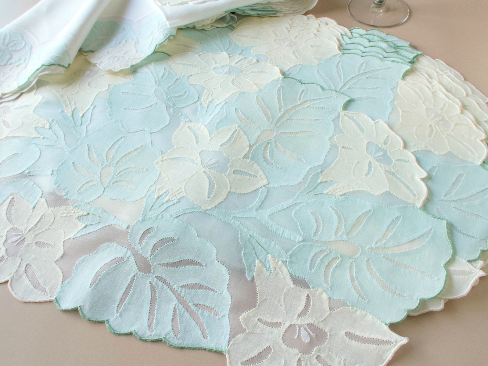 Stack of vintage Madeira organdy placemats showing dimensional hand embroidered flowers and leaves with scalloped edges in pastel tones.