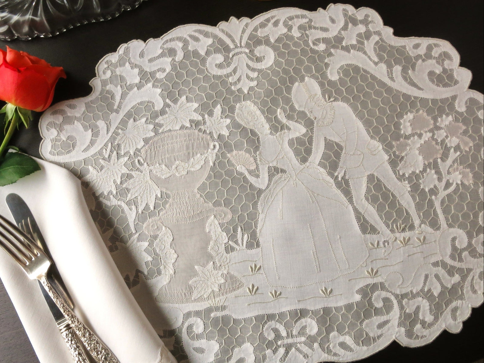 Lace placemat with decorative design on a table setting with cutlery and a rose.