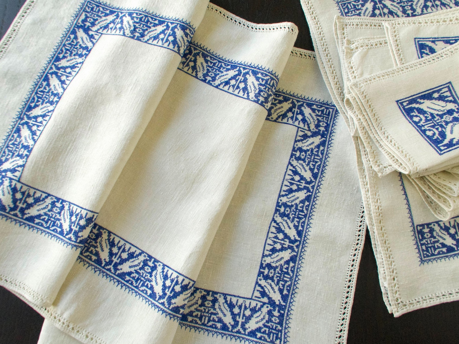 Folded table runner with blue Assisi embroidery and drawnwork edges, beside placemats and napkins from the same set.