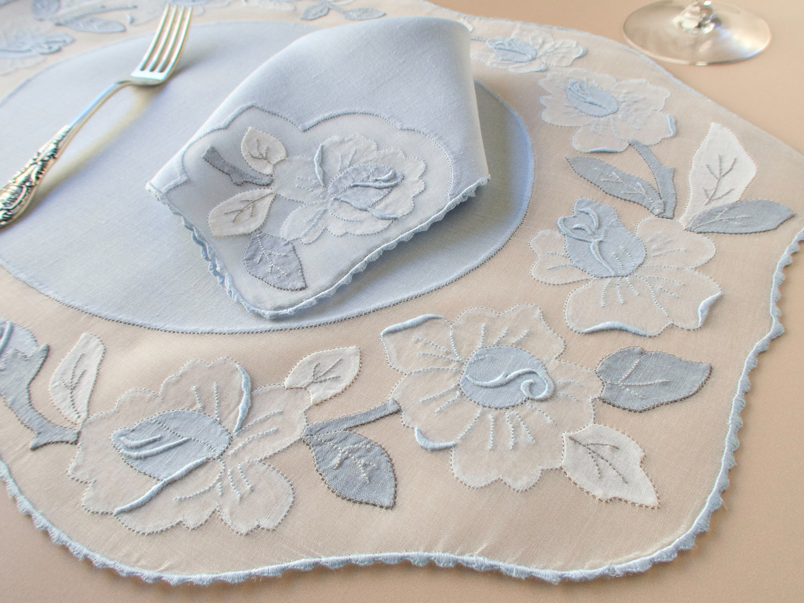 Close-up of vintage Madeira pale blue organdy placemat with large appliqué rose and leaf border in two-tone blue and white, folded Irish linen napkin with matching rose motif, silver cutlery and wine glass on beige background.