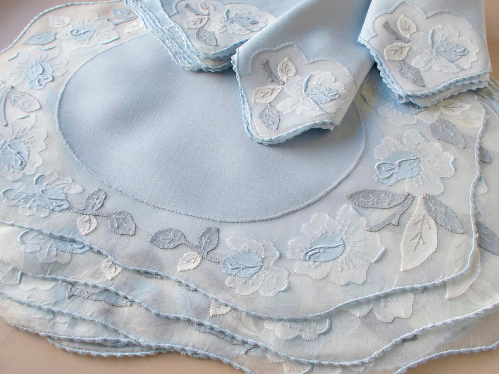 Stack of vintage Madeira pale blue organdy placemats with white and blue appliqué rose border, folded napkins showing set of 8, beige background.