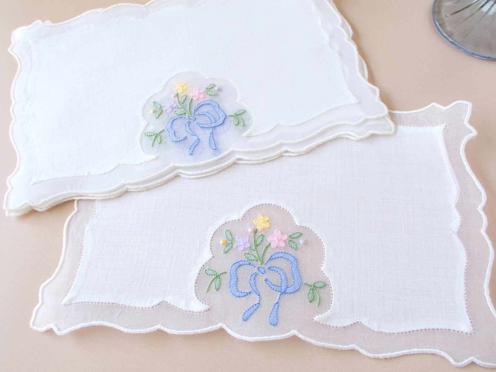 Vintage Madeira cocktail napkins stacked on a beige surface, showing blue ribbon bow embroidery with small flower sprays in yellow and pink on white linen, scalloped organdy appliqué center panel and scalloped outer border.
