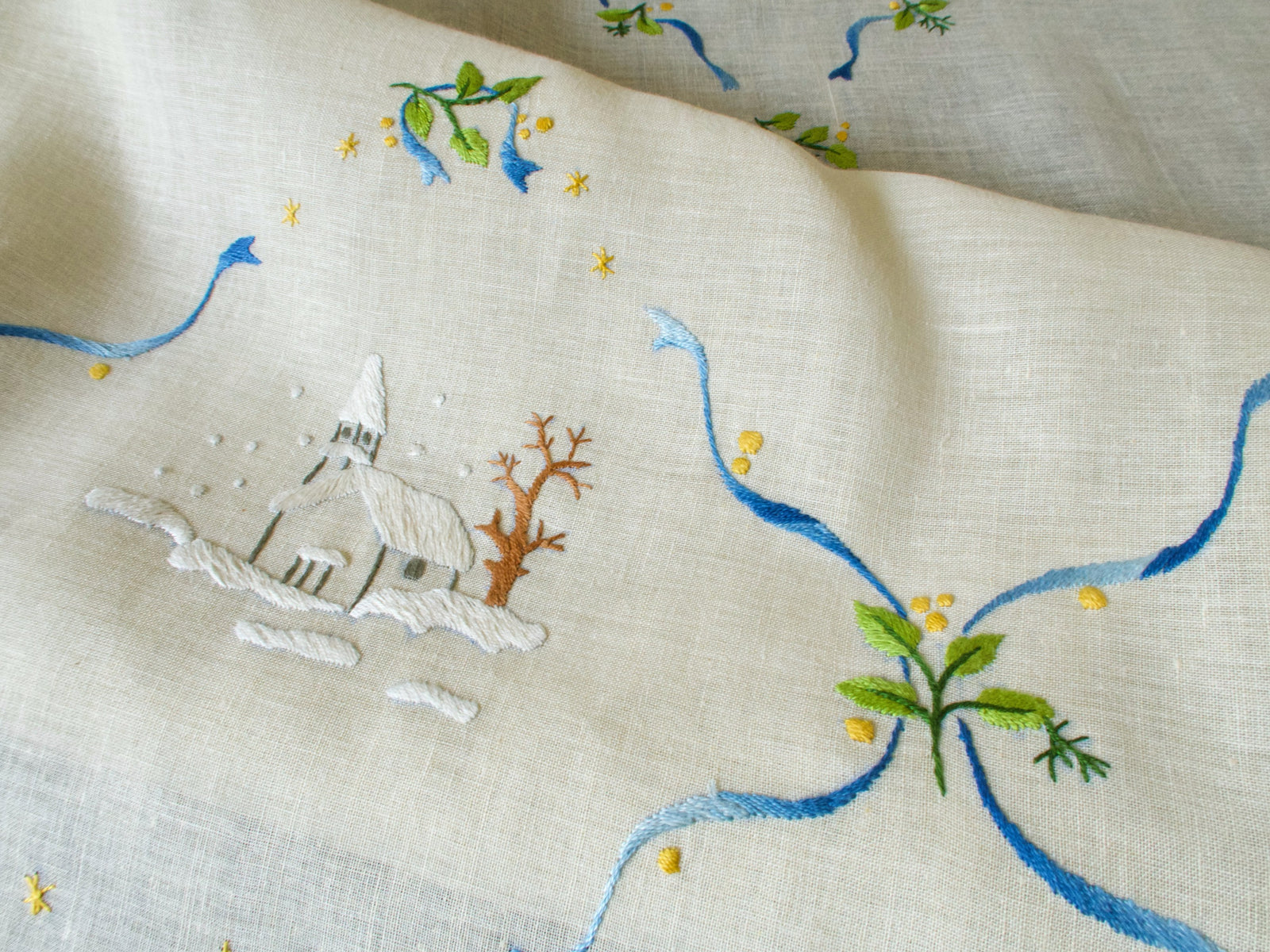 Close-up of embroidered church and trees on vintage linen Christmas tablecloth, showing glowing windows and snow details.