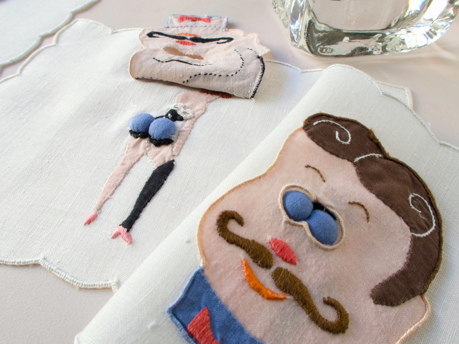 Vintage Madeira peek-a-boo cocktail napkins featuring embroidered men’s faces with blue padded ‘eyes’ that lift to reveal playful pin-up figures underneath. Close-up view showing hand embroidery and dimensional trapunto detail on linen.