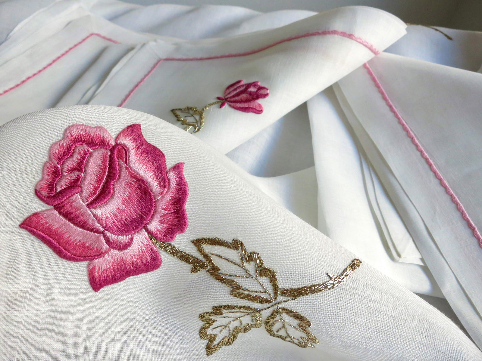 Vintage European cotton batiste tablecloth draped and folded showing hand embroidered pink rose with gold leaf detail on semi-sheer fabric