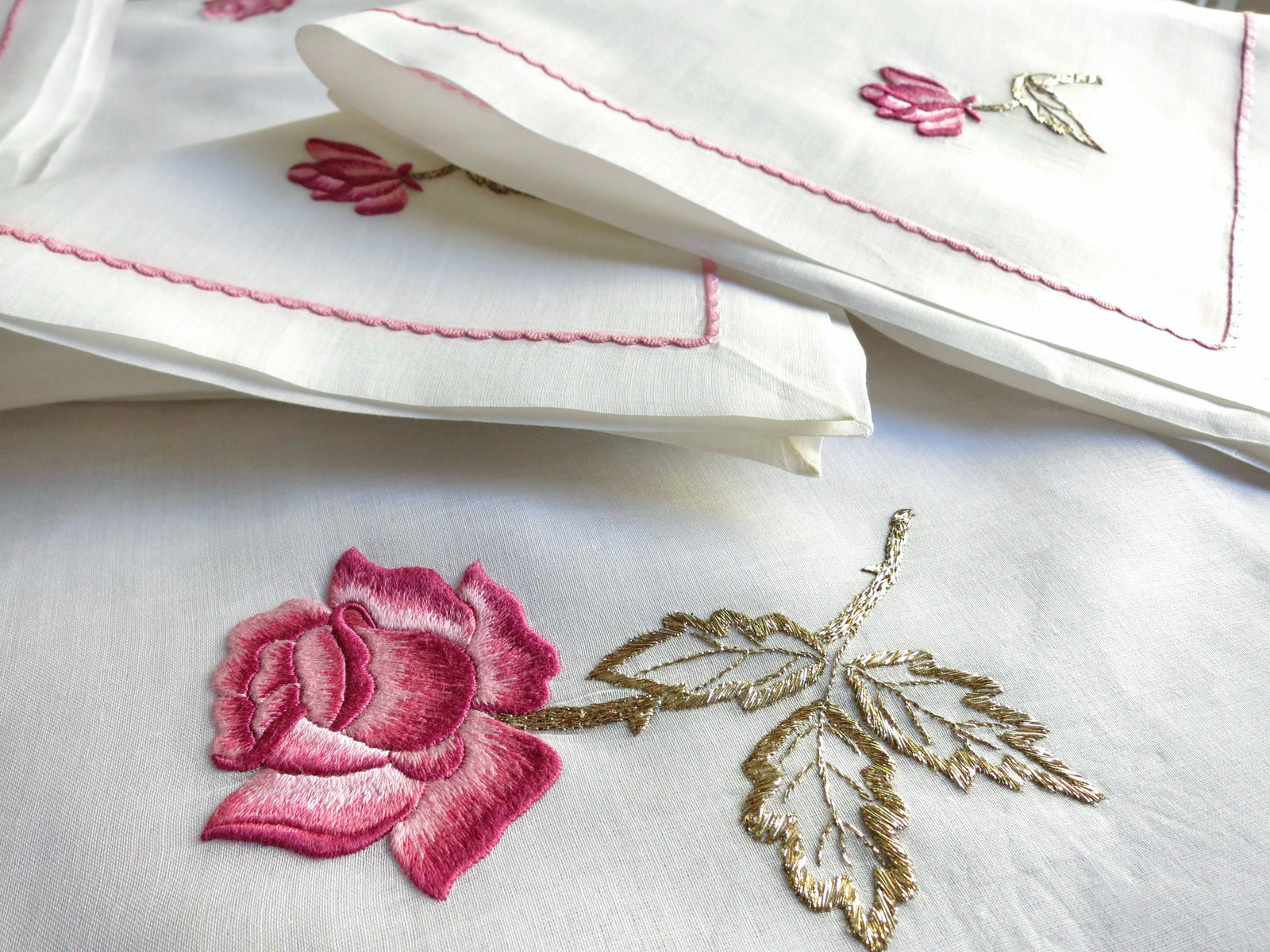 Close-up of vintage European semi-sheer cotton batiste tablecloth hand embroidered with a large pink rose and gold-accented leaves, photographed over white cloth